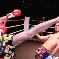 A fighter's leg swings high in a Muay Thai match. The opponent's flinching reflex is evident as he attempts to block the powerful kick.