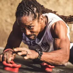 Man with dreadlocks on a climbing wall, his forearm punch showing determination as he grips the holds.
