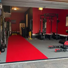 A fully equipped garage gym with a red carpet runner, weight racks, a punching bag, and a "Stronger 40" banner, ready for some serious garage boxing.