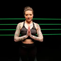 A tattooed woman in athletic wear holds her hands in prayer pose, ready for yoga for boxers.