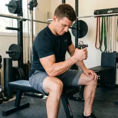 Man in a home gym using hand grippers for forearms. Weights, resistance bands, and a squat rack are visible.