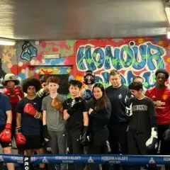 A group of young boxers, some wearing gloves, stand in a boxing ring. One boxer is in the foreground, ready to run.