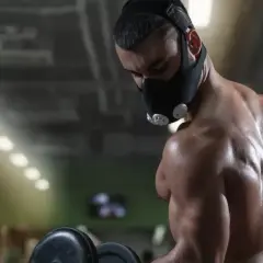 A muscular man performs dumbbell curls in a gym, wearing an oxygen mask for altitude training.