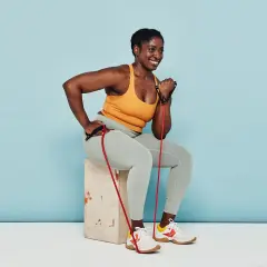 Woman smiles while performing arm strength exercises with a resistance band, seated on a box.