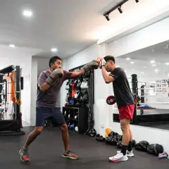 Two men practice boxing conditioning exercises in a gym. One man throws a punch while the other holds pads.