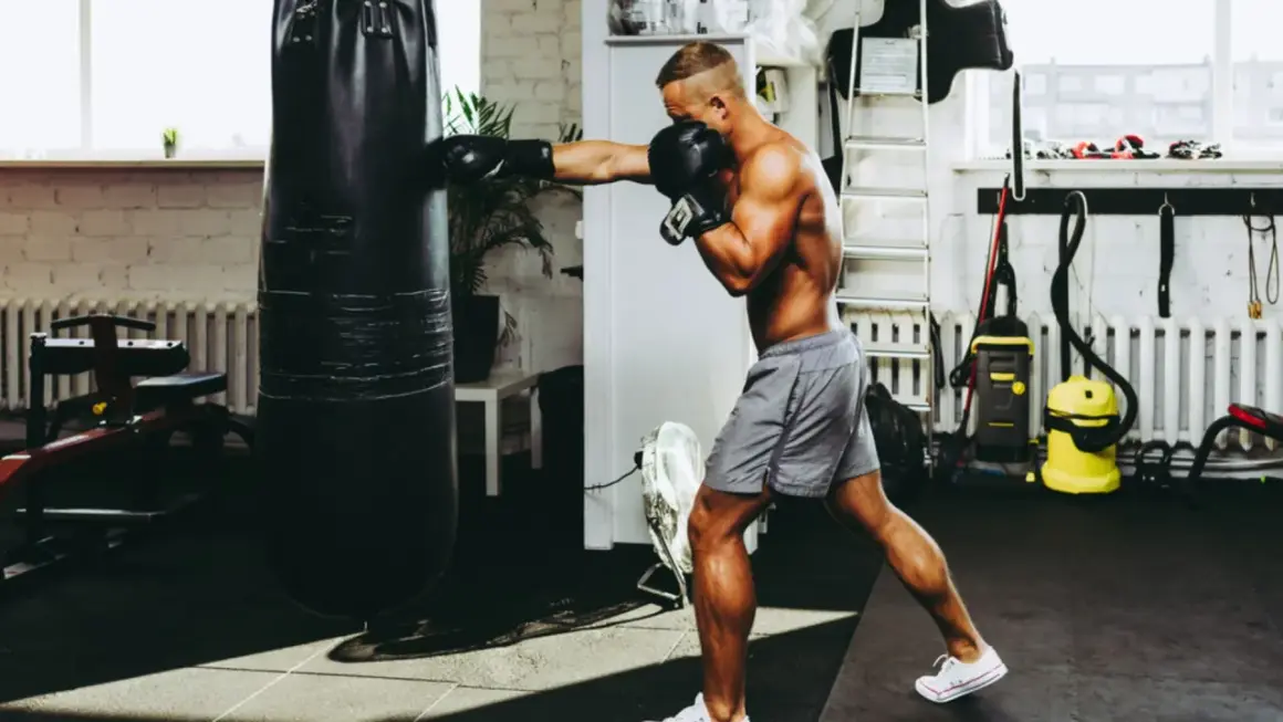 A muscular man punches a heavy bag in a gym, proving that boxing builds muscle.
