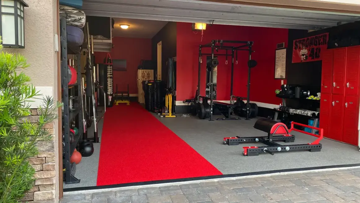 A fully equipped garage gym with a red carpet runner, weight racks, a punching bag, and a "Stronger 40" banner, ready for some serious garage boxing.