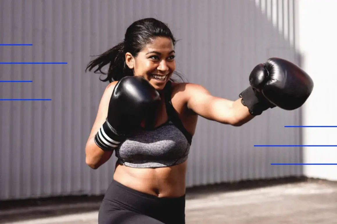 A woman with a bright smile throws a punch during her women boxing exercises. She wears black boxing gloves and athletic wear.