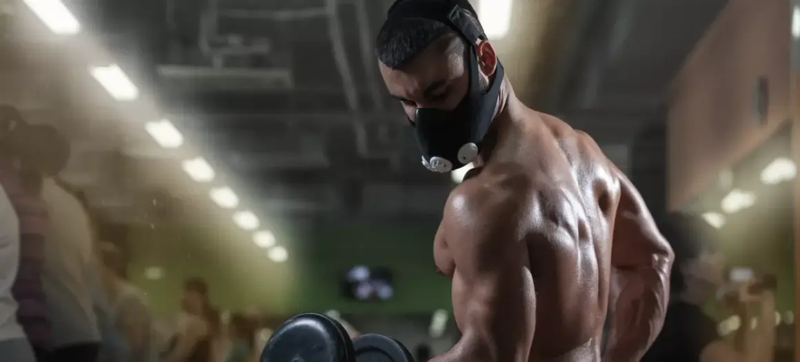 A muscular man performs dumbbell curls in a gym, wearing an oxygen mask for altitude training.