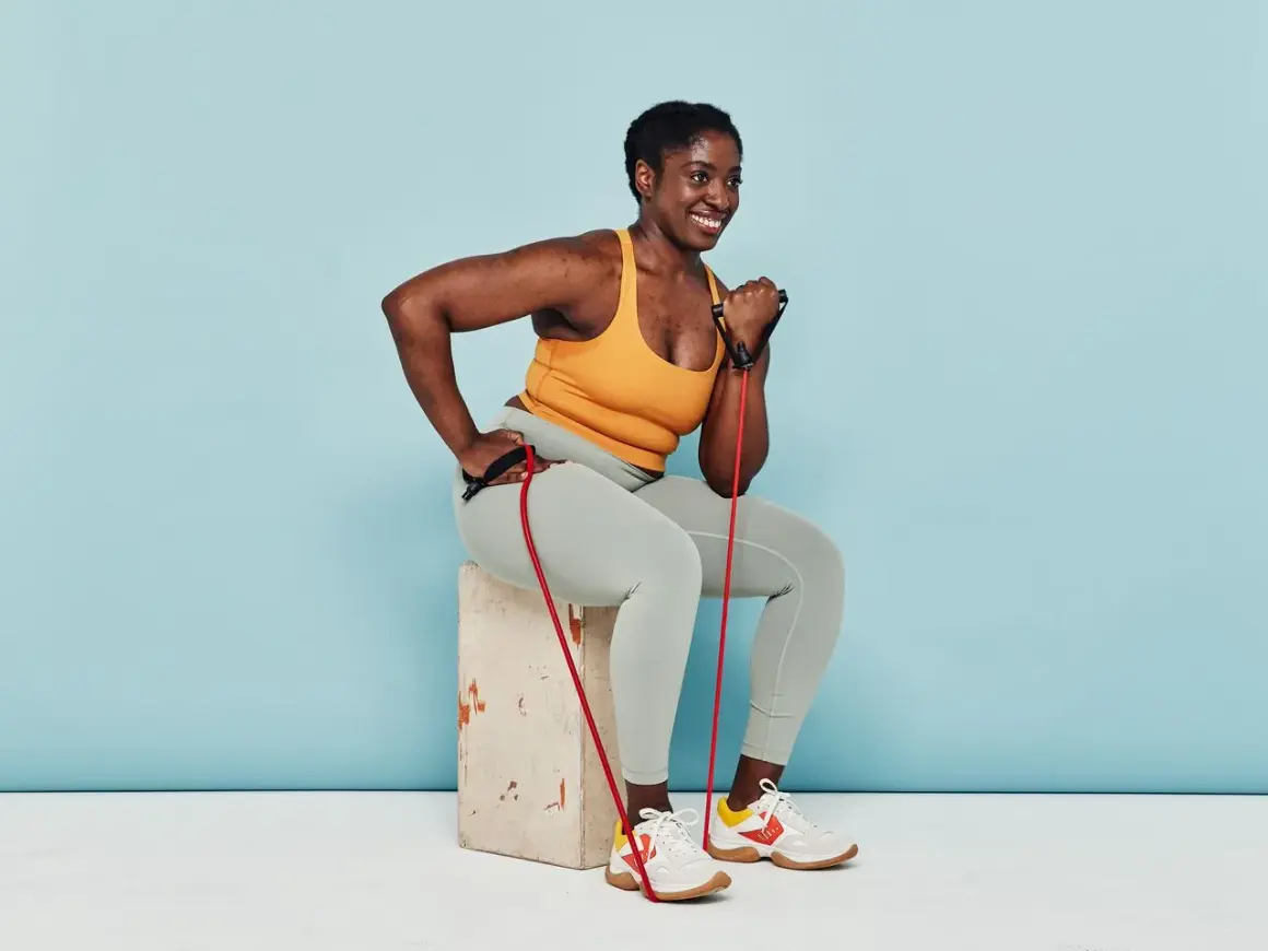 Woman smiles while performing arm strength exercises with a resistance band, seated on a box.