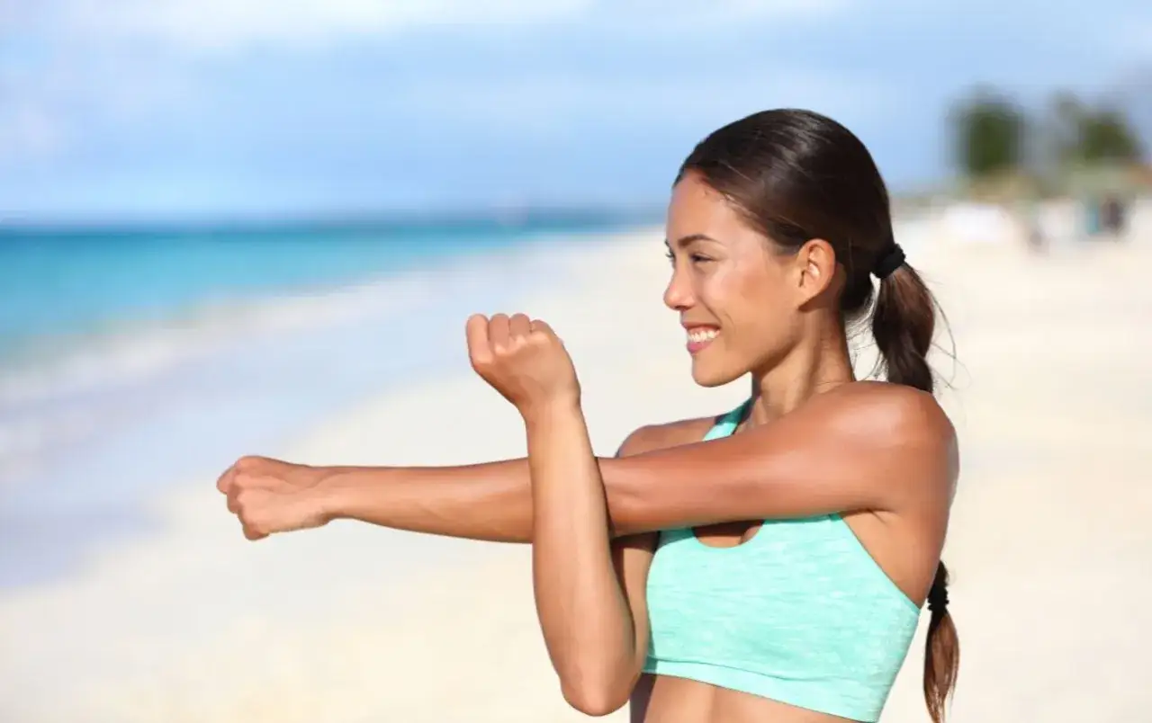 Woman smiling while doing upper body stretches on a beach with the ocean in the background.