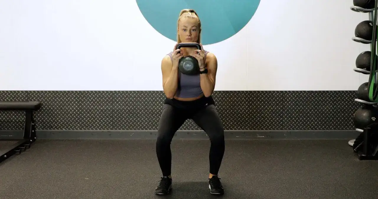 Woman performing pulsing squats with a kettlebell in a gym.