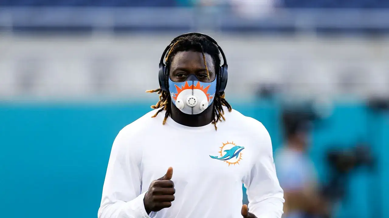A football player wearing headphones and a high elevation mask with the Miami Dolphins logo runs on a field, giving a thumbs up.