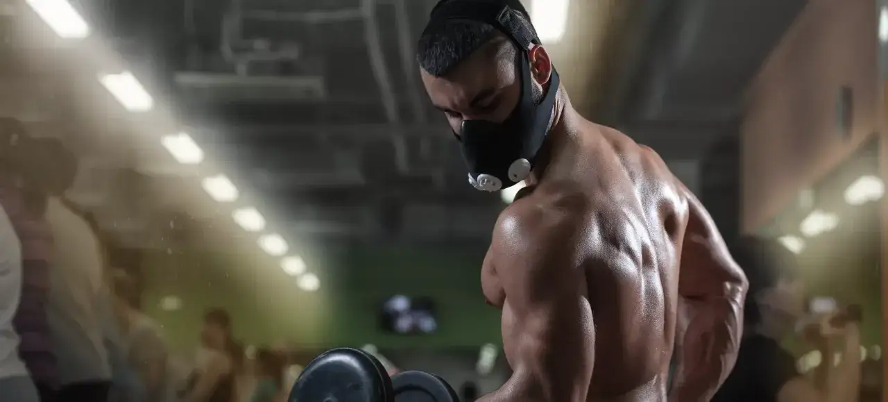 A muscular man performs dumbbell curls while wearing an oxygen mask, pushing his limits during intense oxygen mask training in a gym.