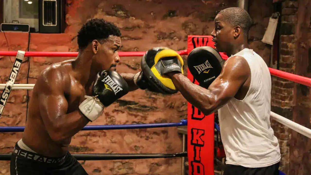 A determined Jacobs boxer trains intensely, sweat glistening as he throws a punch at pads held by his coach in a gritty boxing ring.